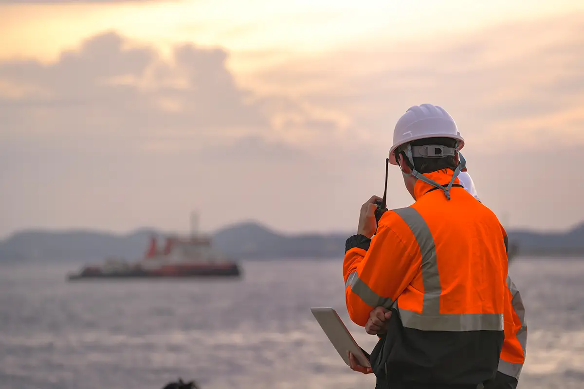 Offshore energy worker in a high-visibility orange jacket and white hard hat using a tablet or radio device, with marine vessels visible in the background at dusk